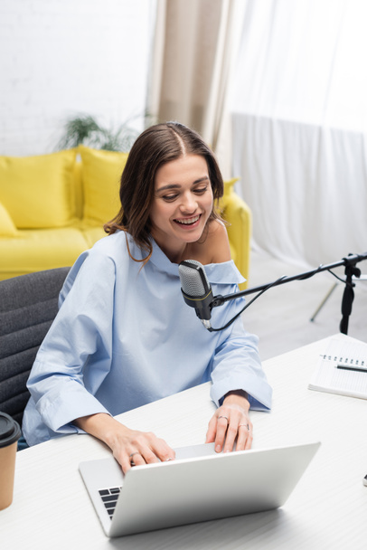 Carefree brunette broadcaster using laptop near microphone during stream, near coffee to go and notebook on table while sitting in podcast studio  - Photo, Image