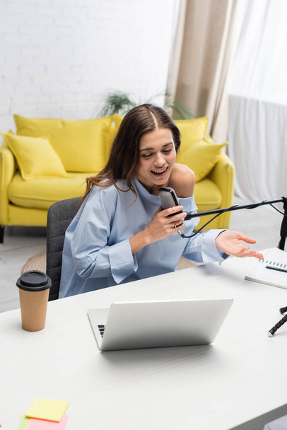 Positive and brunette podcaster talking at microphone, gesturing and looking at laptop near coffee to go and notebook on table during stream in studio  - Foto, Imagen