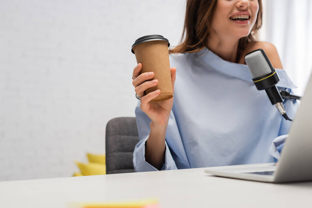 Обрезанный вид веселый и брюнетка подкастер держа кофе, чтобы пойти и говорить на микрофоне рядом размытый ноутбук на столе во время потока в студии  - Фото, изображение