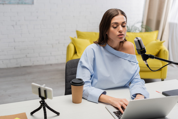 Brunette broadcaster in blouse using laptop near microphone, smartphone on tripod, takeaway coffee and notebook on table during stream  in studio  - Foto, Imagem