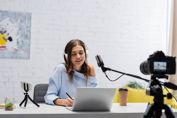 Smiling brunette podcaster in headphones using laptop and holding pen near microphone and digital camera near table during stream in studio  - Foto, Imagem