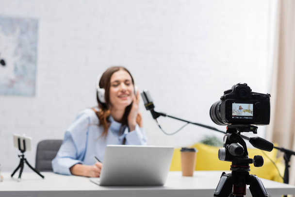 Digital camera near blurred brunette broadcaster in headphones talking during stream near laptop and smartphone on table in podcast studio  - Foto, Imagem