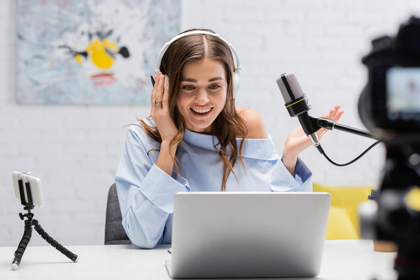 Zorgeloos brunette podcaster in hoofdtelefoon praten en kijken naar laptop in de buurt van microfoon en wazig digitale camera tijdens stream in de studio  - Foto, afbeelding