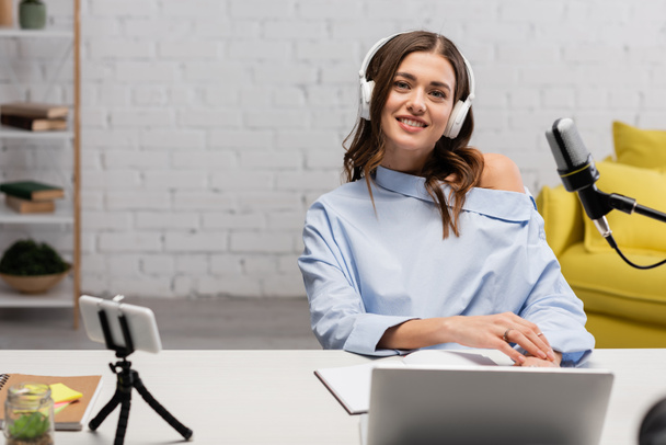 Lächelnde brünette Moderatorin in blauer Bluse und kabellosen Kopfhörern, die in die Kamera neben Mikrofon, Gadgets und Notizbüchern im Studio blickt  - Foto, Bild
