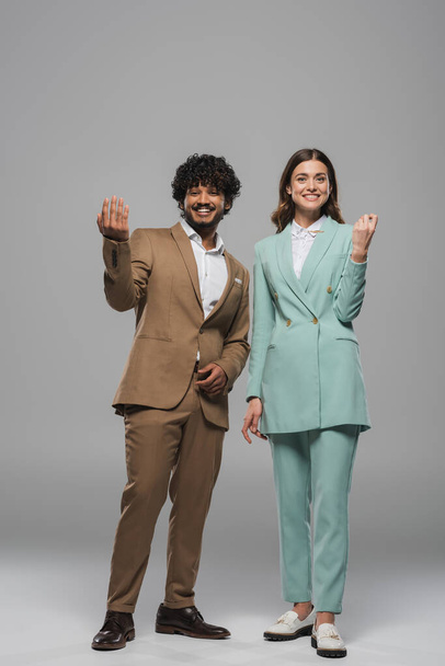 Full length of positive and multiethnic event hosts in formal wear gesturing and looking at camera while standing together on grey background  - Foto, Bild