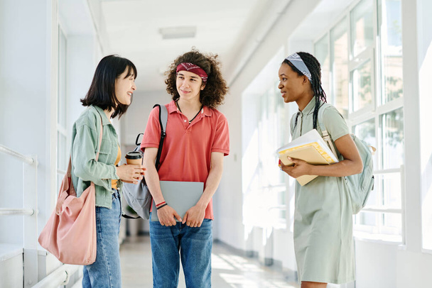 Мультиэтническая группа студентов разговаривает друг с другом во время перерыва стоя в коридоре школьного здания - Фото, изображение