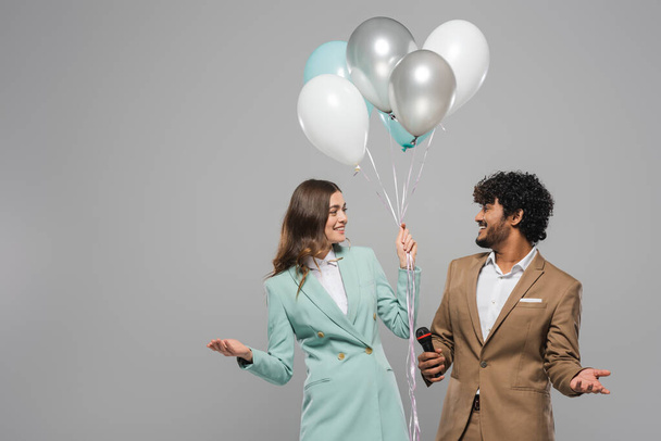 Smiling multiethnic event hosts in formal wear holding microphone and festive balloons while looking at each other and standing isolated on grey   - Photo, Image