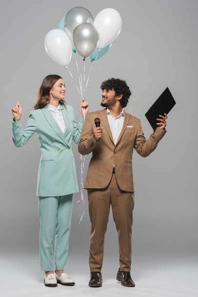 Full length of smiling indian event host in formal wear holding microphone and clipboard near brunette colleague with balloons on grey background  - Fotoğraf, Görsel