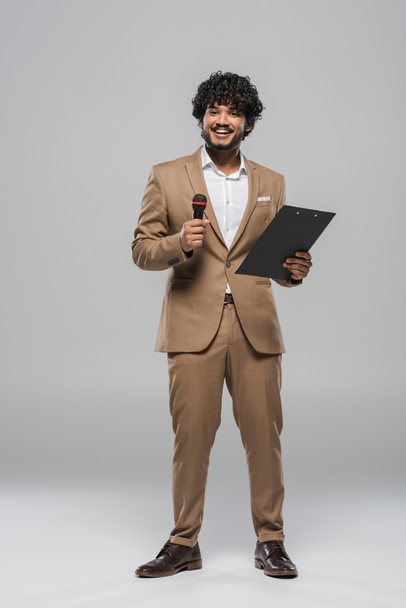 Full length of smiling indian event host in brown formal wear holding microphone and clipboard while standing and smiling on grey background  - Photo, Image