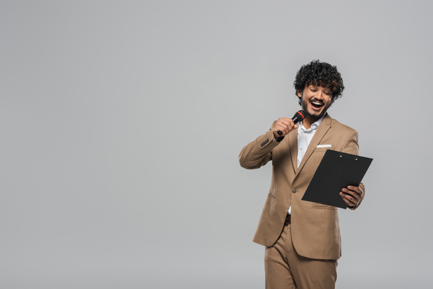 Young and overjoyed indian event host in suit talking at microphone and looking at clipboard during party while standing isolated on grey with copy space - Foto, Imagem