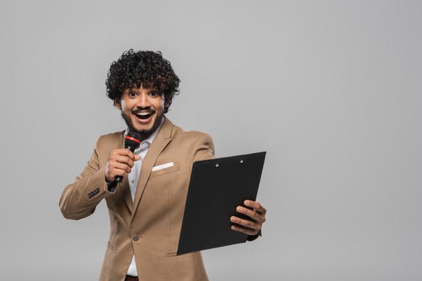 Cheerful and young indian host of event with curly hair standing in formal wear talking at microphone and holding clipboard during party isolated on grey   - Photo, Image