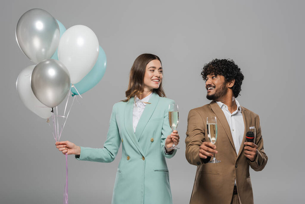 Positive interracial event hosts in formal wear with microphone and festive balloons holding champagne glasses and looking at each other isolated on grey   - Photo, Image