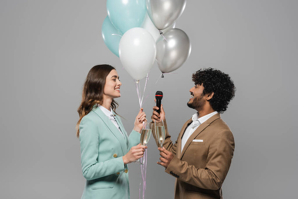 Unbekümmerte Gastgeber in Anzügen mit festlichen Luftballons, Mikrofon und Champagnergläsern während der Party, während sie isoliert auf grau stehen   - Foto, Bild