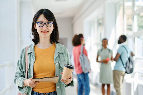 Portrait of Asian college student in eyeglasses drinking coffee and smiling at camera while standing at corridor - Photo, image