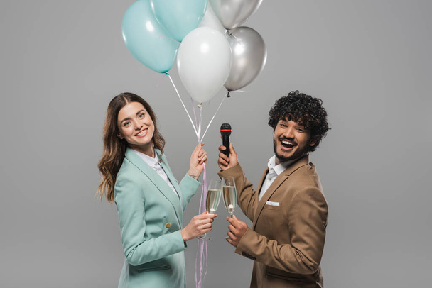 Carefree multiethnic event hosts in formal wear holding festive balloons, microphone and glasses of champagne while looking at camera together isolated on grey   - Foto, afbeelding