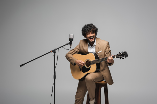 Fröhlicher indischer Moderator in formeller Kleidung spielt Akustikgitarre in der Nähe des Mikrofons, während er auf einem Stuhl sitzt und die Kamera isoliert auf grau anschaut   - Foto, Bild