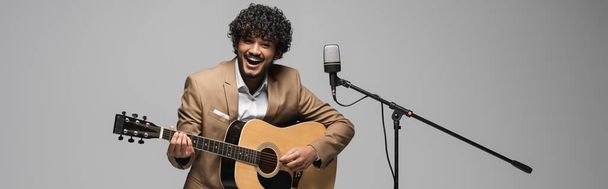 Smiling young indian event host playing acoustic guitar and singing in microphone and looking at camera during performance isolated on grey, banner  - Foto, imagen