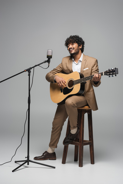 Smiling indian event host in formal wear playing acoustic guitar and singing in microphone in stand while sitting on chair on grey background  - Foto, immagini