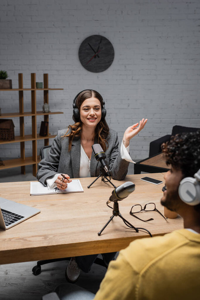 Smiling podcast host in wireless headphones talking to blurred indian guest near notebook, pen, glasses, microphones and devices with copy space during stream in studio  - Foto, imagen
