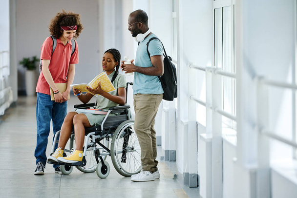 Chica africana con discapacidad abriendo libro de texto y discutiéndolo con sus compañeros de clase mientras están de pie en el pasillo después de la lección - Foto, Imagen