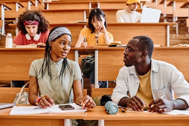 African students talking to each other while sitting at desk at auditorium during lecture - 写真・画像
