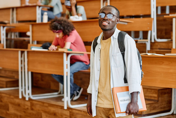 アフリカ人学生の眼鏡の肖像バックパックは、大学の講堂に立ってカメラで微笑んだ - 写真・画像