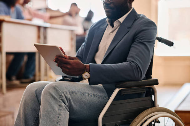 Nahaufnahme eines afrikanischen Lehrers mit Behinderung, der im Rollstuhl sitzt und während des Vortrags ein digitales Tablet benutzt - Foto, Bild
