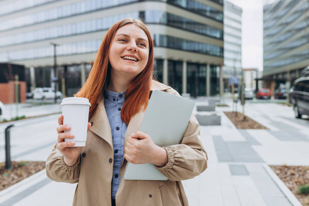 Kaukasische rothaarige Geschäftsfrau in modischer Bürokleidung posiert draußen mit einem Laptop und einer Tasse Kaffee oder Tee. Business on the go Konzept. - Foto, Bild