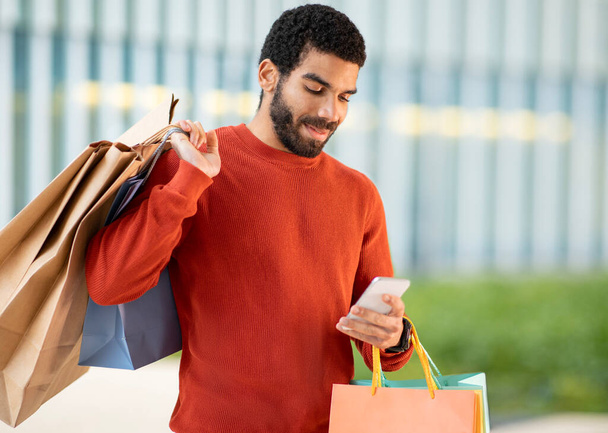 Shopping App. Contented Arabic Male Customer Using Phone Purchasing New Clothes Online In Application Standing Holding Paper Shopper Bags Outside. Gadgets And Ecommerce Concept - 写真・画像