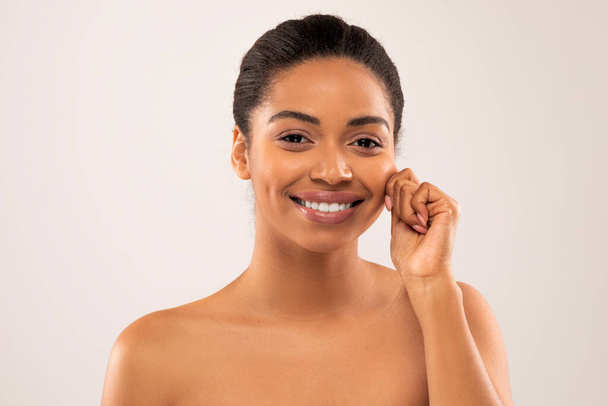 Alegre feliz sonriente atractiva desnuda milenaria afroamericana mujer tocando su mejilla y sonriendo en el fondo del estudio, mostrando la piel brillante suave joven, primer plano - Foto, Imagen
