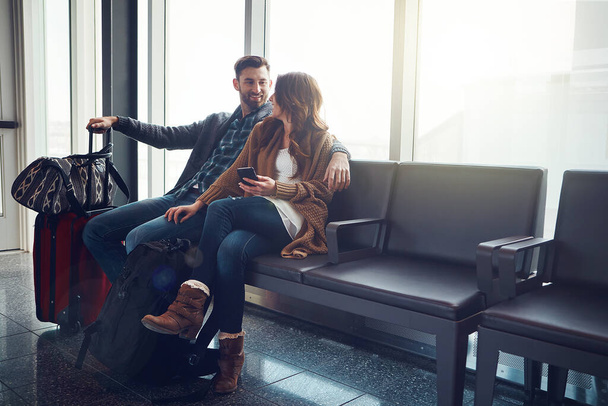 Uçak biraz daha bekleyebilir. Havaalanında bavullarıyla oturan genç bir çift birbirlerine bakıp gülümsüyor. - Fotoğraf, Görsel