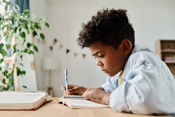 Menino africano do ensino fundamental sentado à mesa com livro e caderno e fazendo lição de casa em casa - Foto, Imagem
