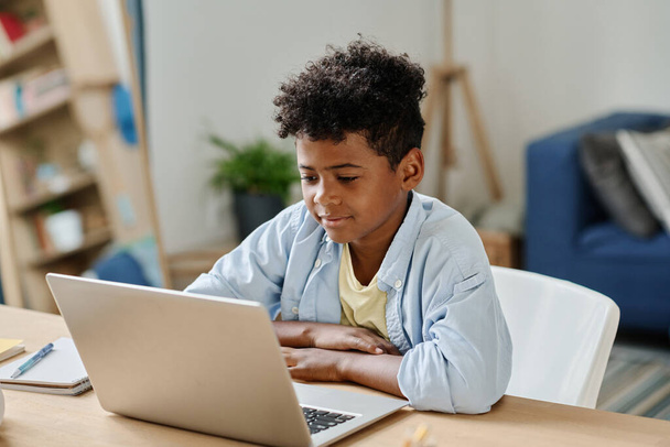 Afrikalı okul çocuğu odasında oturmuş dizüstü bilgisayarını izliyor ve internetten ders çalışıyor. - Fotoğraf, Görsel