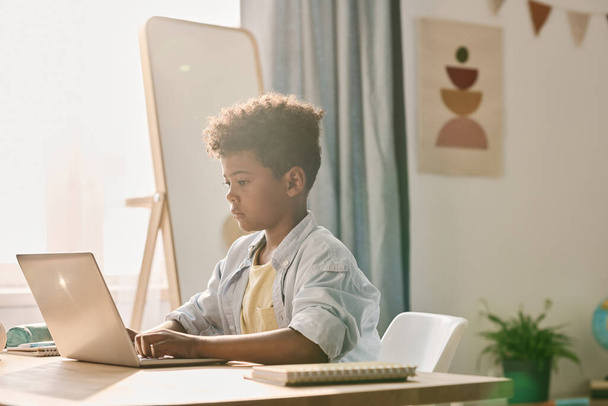 Africain écolier dactylographier sur ordinateur portable pendant la leçon en ligne tout en étant assis à la table dans sa chambre - Photo, image