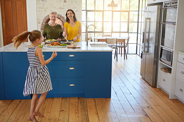 Aufgeregt, das beliebte Familienrezept auszuprobieren. ein kleines Mädchen tanzt, während Mutter und Großmutter im Hintergrund kochen - Foto, Bild
