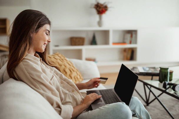 Positive Millennial europäischen weiblichen Eingabe am Computer, Kreditkarte verwenden, genießen Online-Shopping im Wohnzimmer Interieur. Checken Sie Finanzen, Geschäft zu Hause, Gadget für Finanzen und Geldkontrolle - Foto, Bild