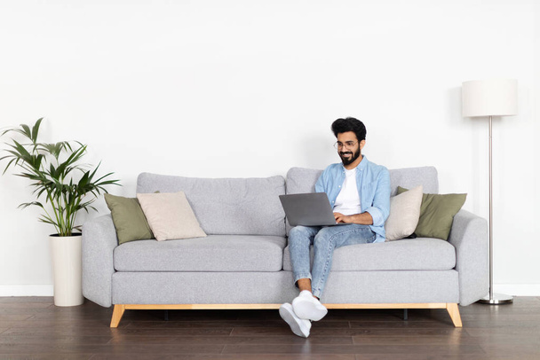 Freelance. Cheerful millennial indian man in casual outfit wearing eyeglasses sitting on sofa over white wall, typing on laptop keyboard and smiling, guy digital nomad working from home, copy space - Foto, afbeelding