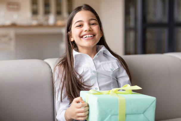 Ευτυχισμένο κορίτσι της Μέσης Ανατολής που κρατά τυλιγμένο κουτί δώρου χαμογελώντας στην κάμερα που κάθεται στον καναπέ στο σπίτι. Preteen παιδί γιορτάζει γενέθλια απόσπαση διακοπών με παραδοθεί παρόν. Επιλογή εστίασης - Φωτογραφία, εικόνα