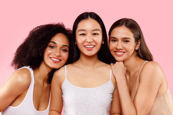 Vielfalt und Schönheitskonzept. Porträt glücklicher drei multiethnischer Damen, die vor rosa Hintergrund posieren und lächelnd in die Kamera lächeln, Studioaufnahme - Foto, Bild