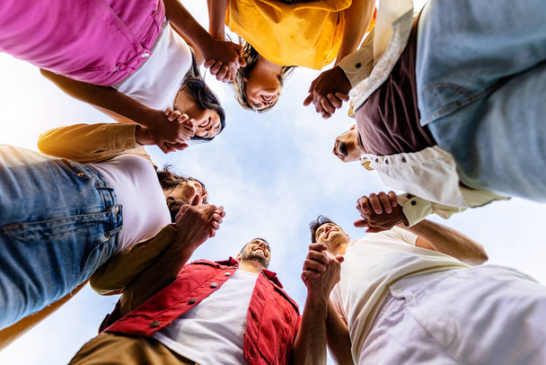 Niski kąt widzenia zjednoczonej grupy młodych ludzi w kręgu trzymających się za ręce. Koncepcja wspólnoty, wsparcia i pracy zespołowej z tysiącletnimi ludźmi ukazującymi jedność na świeżym powietrzu. - Zdjęcie, obraz