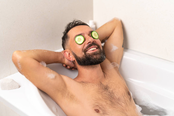 Bearded man with cucumber on front of his eyes relaxing in bath with soapsuds at home - Фото, изображение