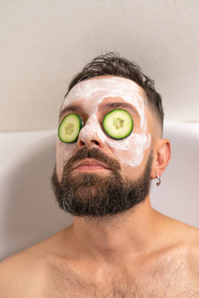 Γενειοφόρος άνδρας με αγγούρι μπροστά στα μάτια του και λευκή μάσκα στο πρόσωπο χαλαρώνει στο μπάνιο στο σπίτι - Φωτογραφία, εικόνα