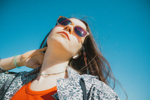 Madrid Spain April 16 2023 Stylish fashion pretty white young woman with long brown hair in wind Female portrait against blue sky Free-spirited millennial generation girl in sunglasses leopard jacket - Photo, image
