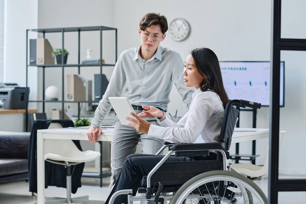 Mujer de negocios asiática con discapacidad apuntando a la tableta digital y discutiendo informe en línea con su colega en la oficina - Foto, imagen