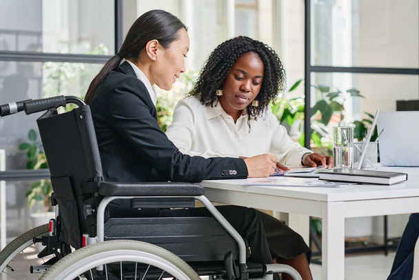 Asian businesswoman in wheelchair discussing business contract with her colleague at table at office - Photo, Image