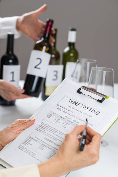 Füllen Sie ein Formular für eine Weinprobe aus. Verkostungsgast bewertet die Eigenschaften eines Rotweins und füllt ein Verkostungsformular aus, um Beobachtungen aufzuzeichnen. - Foto, Bild