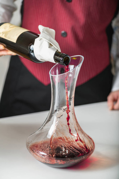 Αποξηραίνοντας το κρασί. Ένας οινοχόος ρίχνει κόκκινο κρασί από ένα μπουκάλι σε μια καράφα, εξηγώντας πώς να αερίσει το κρασί για να βελτιώσει τη γεύση και το άρωμά του. - Φωτογραφία, εικόνα