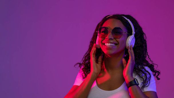 無線電話で音楽を聞くスタイリッシュなサングラスを身に着けている陽気なアフリカ系アメリカ人女性,紫のスタジオの背景にネオンで立って幸せな黒の女性を笑顔,スペースをコピー - 写真・画像