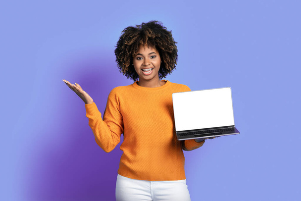 Amazed happy young black woman with bushy hair in casual outfit holding computer laptop with white blank screen and gesturing on purple studio background, showing great online deal, mockup - Фото, зображення