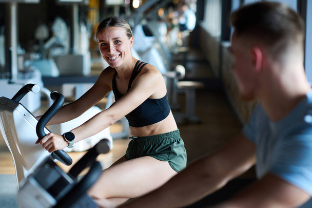 Junge Frau lächelt ihrem Freund zu, während sie gemeinsam auf Fahrrad-Trainingsgerät im Fitnessstudio trainieren - Foto, Bild
